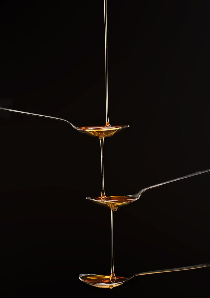 team-02 Artistic close-up of golden honey streaming from three aligned spoons on a black background.
