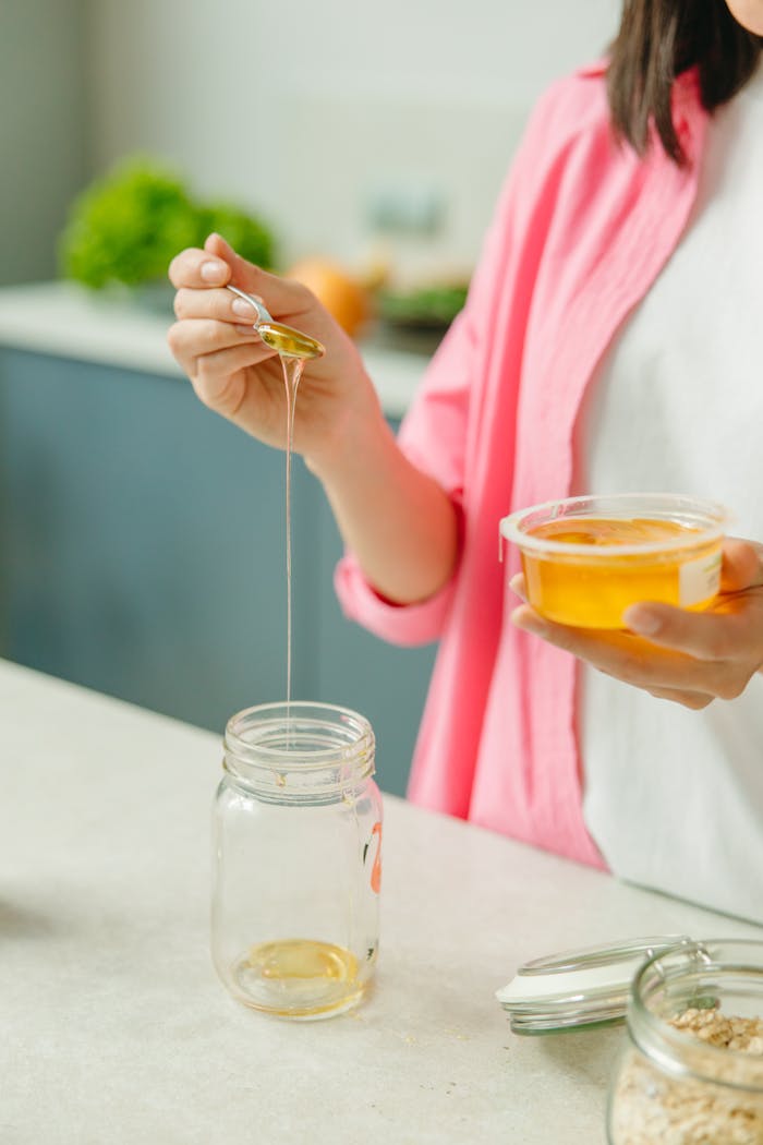 who-we-are Close-up of a woman pouring honey from a spoon into a glass jar in the kitchen, healthy lifestyle